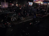 The square is filled with cars, motorcycles, and barrels with fires burning in them in 1985-A.