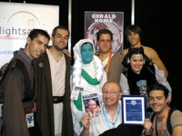 Gerald Home recibe la placa conmemorativa de membresía honorífica de HoloRed Estelar Sevilla junto al Fanzine en el que partició en una entrevista.
